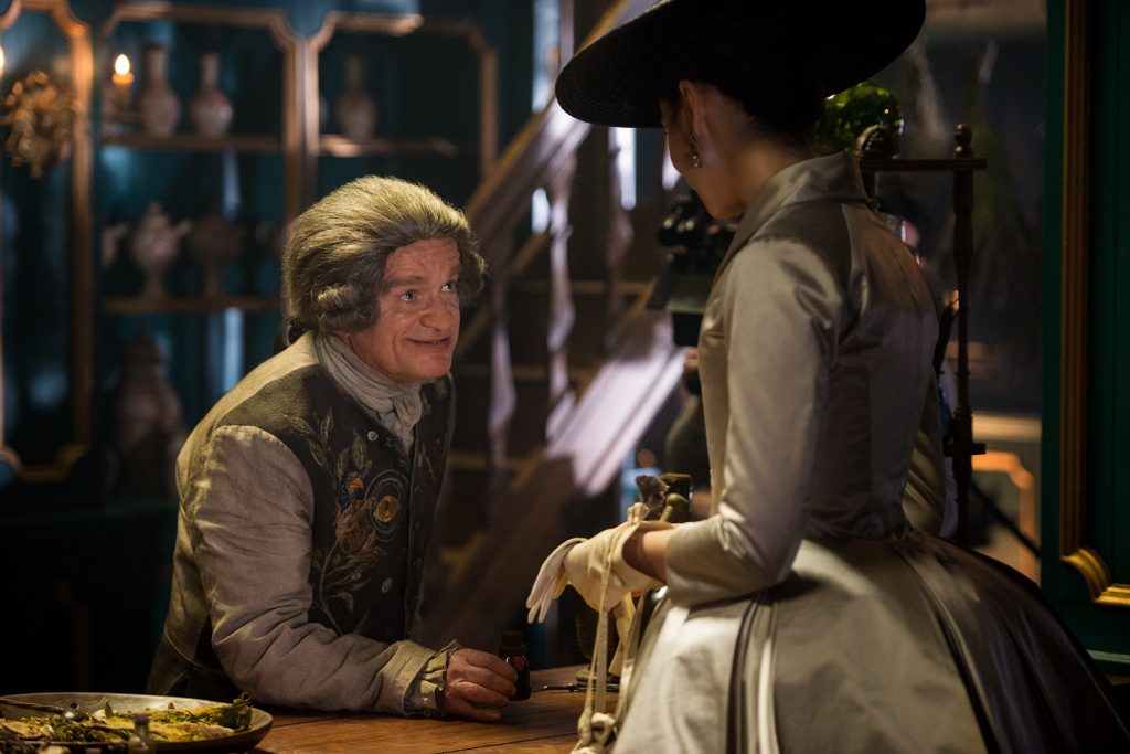 Clearly a man with such fashion sense could never be guilty. (Left, Dominique Pinon as Master Raymond speaking to Claire Fraser on the right.)
