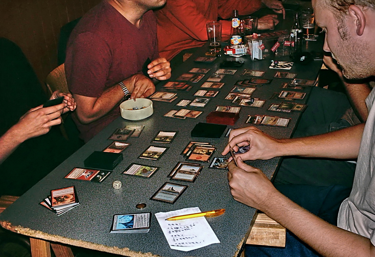 People sit at a table spread with colorfol cards. A session of Magic: The Gathering. [Courtesy Wikimedia Commons]